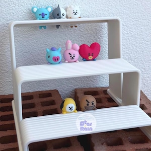 May include: A white, two-tiered display shelf holds several colorful character figurines. The figurines include a blue koala, a gray character with a split face, a white sheep, a blue creature with a heart, a pink bunny, a red heart, a yellow character, and a brown character.