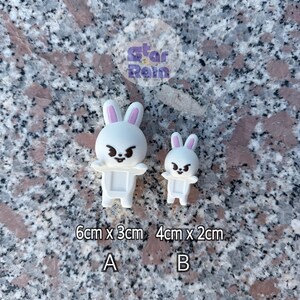 May include: Two white bunny figurines with pink inner ears and angry expressions. The larger bunny measures 6cm x 3cm, and the smaller one is 4cm x 2cm. The text "A" and "B" are below the bunnies.