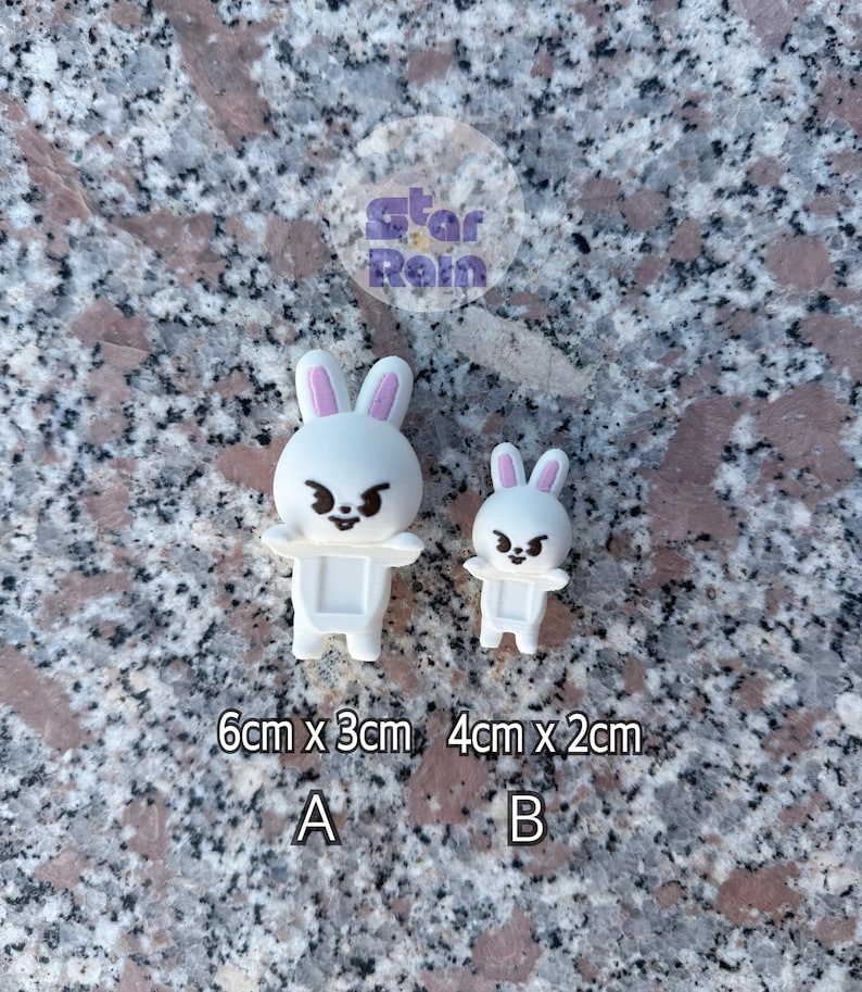 May include: Two white bunny figurines with pink inner ears and angry expressions. The larger bunny measures 6cm x 3cm, and the smaller one is 4cm x 2cm. The text "A" and "B" are below the bunnies.