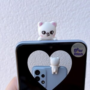 May include: A white, cat-shaped phone charm sits atop a light blue smartphone. The phone's screen displays a heart-shaped cutout revealing a matching phone and charm. The charm has pink accents and a smiling face. The phone has a triple-lens camera.