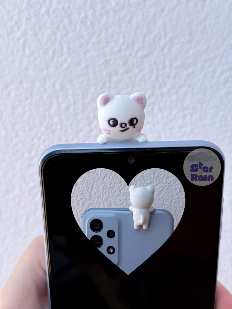 May include: A white, cat-shaped phone charm sits atop a light blue smartphone. The phone's screen displays a heart-shaped cutout revealing a matching phone and charm. The charm has pink accents and a smiling face. The phone has a triple-lens camera.