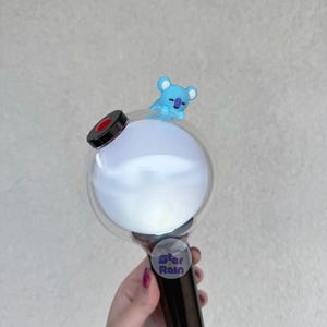 Op de afbeelding: Een zwarte en doorzichtige lichtstaaf met een rond, wit licht en een kleine blauwe koala figuur bovenop. De lichtstaaf heeft een zwarte handgreep en een sticker met de tekst "Star Rain".