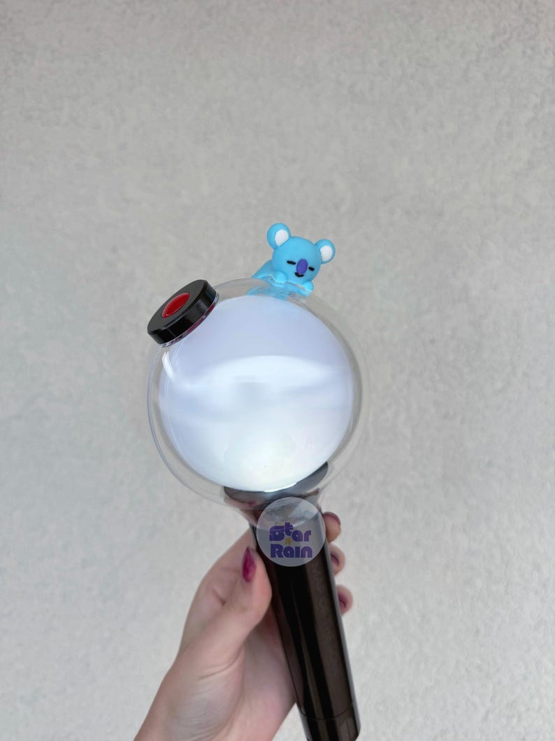 Op de afbeelding: Een zwarte en doorzichtige lichtstaaf met een rond, wit licht en een kleine blauwe koala figuur bovenop. De lichtstaaf heeft een zwarte handgreep en een sticker met de tekst "Star Rain".