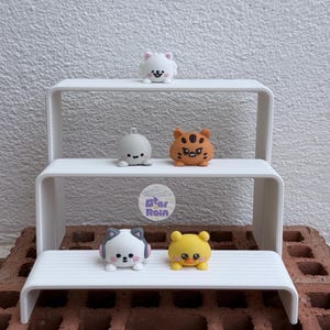 May include: A white three-tiered display stand holding five small animal figurines. The top tier features a white cat. The middle tier holds a gray round creature and an orange tiger. The bottom tier displays a white cat with pink accents and a yellow duck.