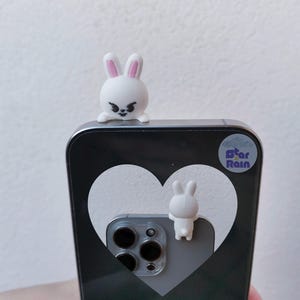 May include: A white phone case with a heart design and two small white bunny figurines. One bunny is perched on top, and the other is attached to the back. The phone case has a black and gray color scheme, with a sticker that says "Star Rain."