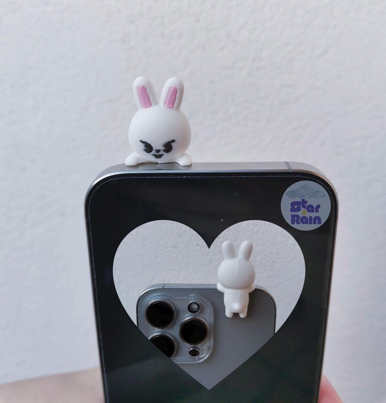 May include: A white phone case with a heart design and two small white bunny figurines. One bunny is perched on top, and the other is attached to the back. The phone case has a black and gray color scheme, with a sticker that says "Star Rain."