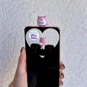 May include: A black smartphone with a gold-colored frame, featuring a pink bear-shaped phone charm. The phone case has a heart-shaped cutout with the text "star Rain". The bear charm is pink with a white belly and a smiling face.