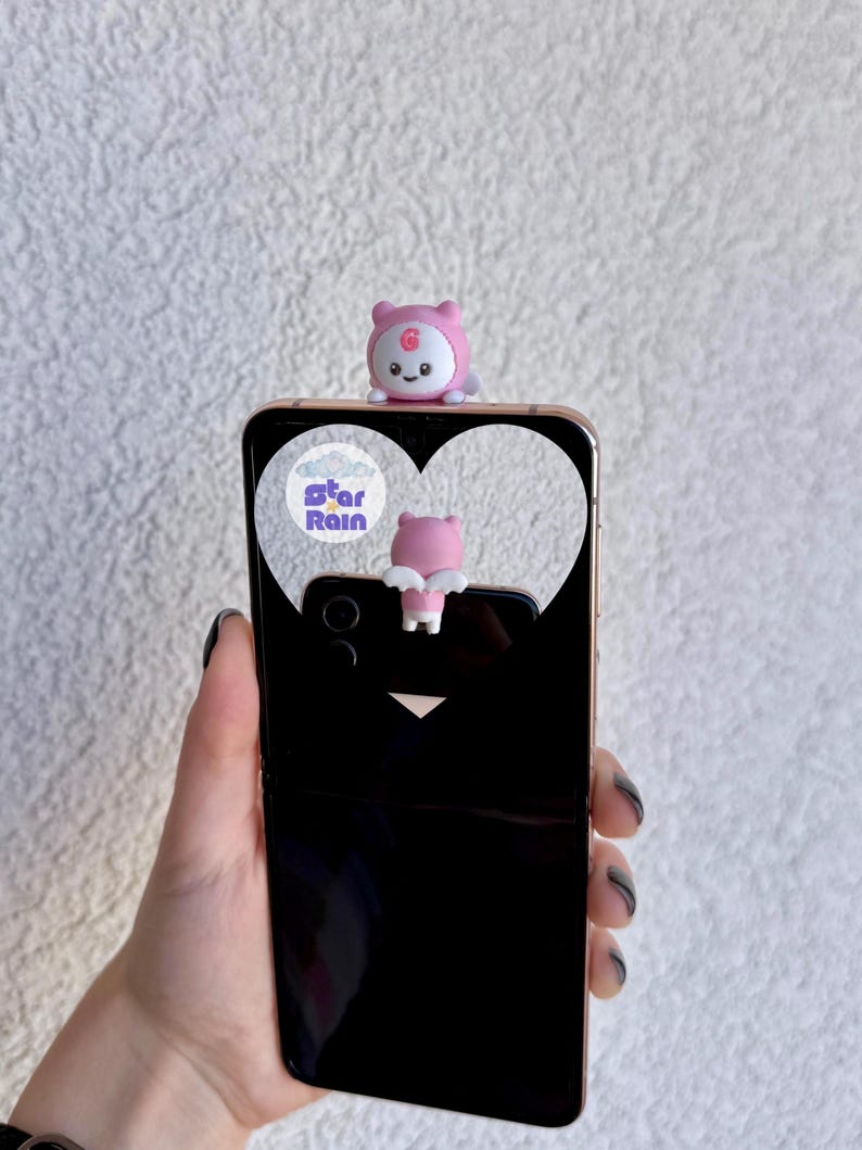 May include: A black smartphone with a gold-colored frame, featuring a pink bear-shaped phone charm. The phone case has a heart-shaped cutout with the text "star Rain". The bear charm is pink with a white belly and a smiling face.