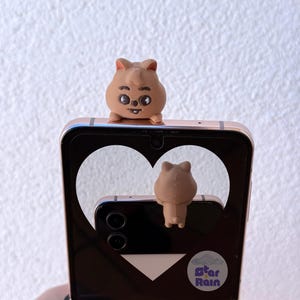 May include: A brown quokka phone grip and a matching quokka phone charm are attached to a gold-colored smartphone. The phone's screen displays a heart-shaped cutout. The phone charm is attached to the back of the phone.