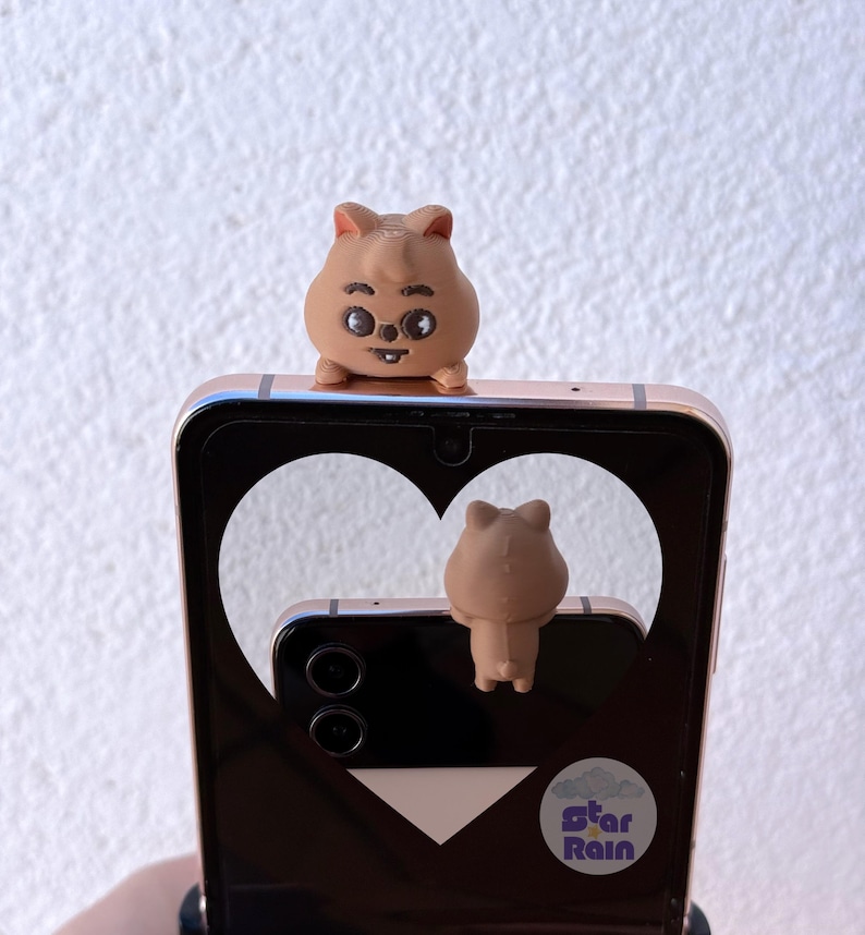 May include: A brown quokka phone grip and a matching quokka phone charm are attached to a gold-colored smartphone. The phone's screen displays a heart-shaped cutout. The phone charm is attached to the back of the phone.