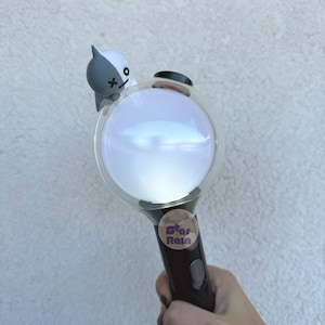 Op de afbeelding: Een zwart-witte lichtstaaf met een heldere, bolvormige kop. Een klein stripfiguur zit bovenop de bol. De handgreep heeft een knop en een sticker met de tekst "Star Rain".
