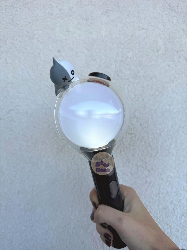 Op de afbeelding: Een zwart-witte lichtstaaf met een heldere, bolvormige kop. Een klein stripfiguur zit bovenop de bol. De handgreep heeft een knop en een sticker met de tekst "Star Rain".