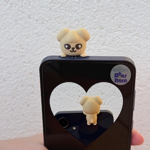 May include: A beige puppy-shaped phone charm sits atop a navy blue phone case. The phone case has a heart-shaped cutout, and the text "Star Rain" is visible. The charm has black eyes and a small pink mouth.