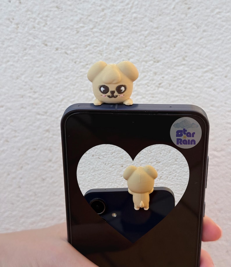 May include: A beige puppy-shaped phone charm sits atop a navy blue phone case. The phone case has a heart-shaped cutout, and the text "Star Rain" is visible. The charm has black eyes and a small pink mouth.