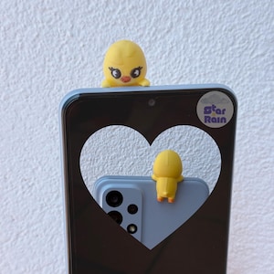 May include: A light blue smartphone case with a heart-shaped cutout, featuring a yellow duck figurine. The case has a camera module and the text "Star Rain". A second yellow duck figurine is on the back of the phone.
