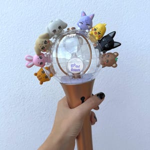 Puede incluir: Una varita de concierto iluminada con un globo transparente y un mango de color bronce. El globo está adornado con pequeñas y coloridas figuras de animales de dibujos animados. El texto "Star Rain" es visible en el globo.