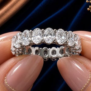 Puede incluir: Un deslumbrante anillo de la eternidad con una fila continua de diamantes de talla ovalada engastados en una banda plateada. Los diamantes brillan, mostrando sus facetas. El anillo se presenta sobre un fondo azul oscuro, resaltando su elegancia.