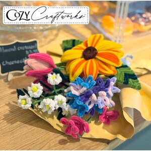 May include: A colorful bouquet of handmade pipe cleaner flowers, including a large yellow and orange sunflower, pink tulips, white daisies, and blue and purple starburst-shaped flowers. The flowers are arranged on a yellow paper and displayed on a wooden surface. The text "COZY Craftworks" is in the top left corner.