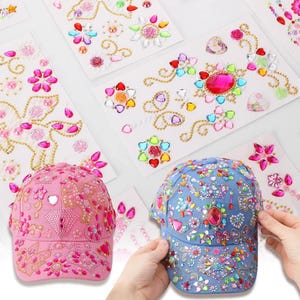 Puede incluir: Gorras de béisbol rosas y azules adornadas con pedrería y gemas de colores. También se ven hojas de pegatinas decorativas con diseños florales y de corazón, con detalles en tonos joya y detalles dorados.