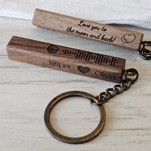 Peut inclure: Deux porte-clés en bois avec des anneaux en métal. L'un porte l'inscription "Love you to the moon and back !" et un cœur. L'autre porte un code Spotify, les noms "Jasper" et "Olivia" et un cœur.