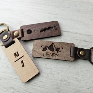 May include: Three wooden keyrings with leather straps and metal rings. One keyring has the initials "M/J", another has a mountain graphic and the name "HENRY", and the third has a music waveform.