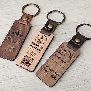 May include: Three wooden keyrings with leather straps and metal rings. One keyring has the text "Mason Family Home" and a mountain graphic. Another says "Whispering Pines Cabin" with a cabin graphic and a QR code. The third says "Rest Relax Recharge".