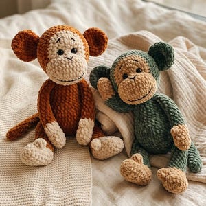 Può includere: Due peluche a forma di scimmia fatti a mano all'uncinetto. Uno è marrone e crema, l'altro verde e beige. Entrambi hanno facce sorridenti, grandi orecchie e sono seduti su una coperta color crema. Questi morbidi giocattoli sono perfetti per i bambini.
