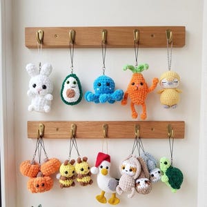 Könnte beinhalten: Eine Sammlung handgefertigter Häkel-Schlüsselanhänger, darunter ein weißes Kaninchen, eine Avocado, ein blauer Oktopus, eine orangefarbene Karotte, ein gelbes Faultier, orangefarbene Kürbisse, eine Biene, eine Ente mit Weihnachtsmütze, ein Faultier und eine grüne Schildkröte.