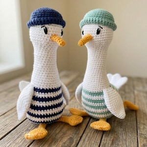 Könnte beinhalten: Zwei gehäkelte Gänse mit weißen Körpern, gelben Schnäbeln und Füßen. Eine Gans trägt eine marineblaue Mütze und ein blau-weiß gestreiftes Shirt, die andere eine grüne Mütze und ein grün-weiß gestreiftes Shirt.