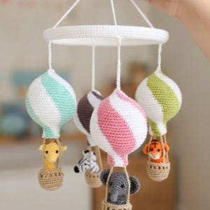 Könnte beinhalten: Handgefertigtes Baby-Mobile aus Häkelarbeit mit fünf Heißluftballons in verschiedenen Farben, darunter Weiß, Rosa, Grün und Braun. Jeder Ballon hat einen Korb mit einem gehäkelten Tier, wie einer Giraffe, einem Zebra, einem Tiger und einem Elefanten.