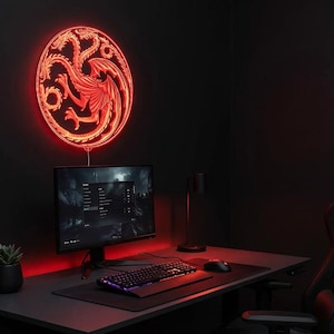 May include: A red neon sign in the shape of a dragon emblem is mounted on a dark wall above a computer desk. The desk has a monitor, keyboard, mouse, and a small potted plant. A gaming chair is in the corner.