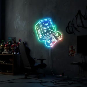 May include: A neon sign in the shape of a retro video game console holding a skateboard, with the letters "BMC" on the console. The sign glows with blue, green, and pink lights, mounted on a gray wall. A black gaming chair is in the foreground.