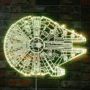 K&ouml;nnte beinhalten: Eine neongr&uuml;ne Millennium Falcon Wandleuchte, die das komplizierte Design des Schiffs zeigt. Die Leuchte ist an einer Backsteinmauer befestigt, was einen auff&auml;lligen Kontrast erzeugt. Der Umriss der Leuchte leuchtet und hebt die Details des Schiffs hervor.