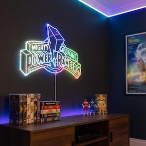 Könnte beinhalten: Neon-Schild mit der Aufschrift "Mighty Morphin Power Rangers" in einem leuchtenden, mehrfarbigen Design. Das Schild ist an einer dunklen Wand über einem Holzschrank mit VHS-Kassetten und Actionfiguren befestigt.
