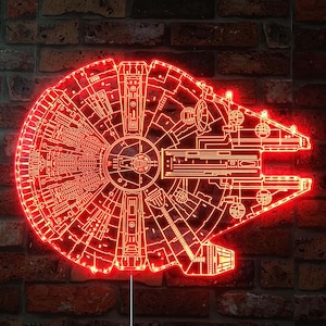 K&ouml;nnte beinhalten: Rotes LED-Licht in Form des Raumschiffs Millennium Falcon. Das Licht zeigt eine detaillierte Umrisszeichnung des Schiffsdesigns mit rotem Leuchten. Das Licht ist an einer Backsteinmauer befestigt.