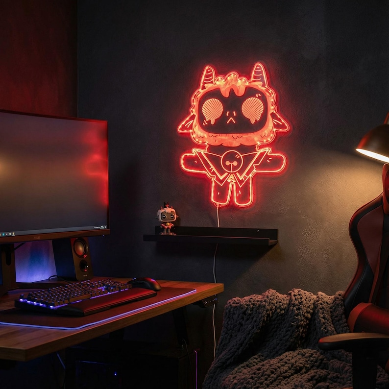 May include: A red neon light in the shape of a cartoon character with horns and a cape is mounted on a dark wall. The light is plugged in and illuminates the room. A computer monitor, keyboard, and mouse are on a wooden desk.
