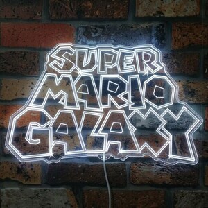 May include: A white neon sign displaying the words "SUPER MARIO GALAXY" on a brick wall. The sign's text is in a distinctive font, illuminated by bright white light. The brick wall features a blend of red, brown, and grey bricks.