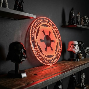 May include: A glowing red Imperial symbol wall light. The circular light features a detailed design with a central emblem and surrounding text. The light is mounted on a dark wall above a wooden shelf with Star Wars figurines.