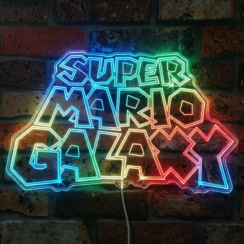 May include: A neon sign displaying "SUPER MARIO GALAXY" in a vibrant, multi-coloured font. The sign's outline glows with a spectrum of colours, including blue, green, yellow, and red. It is fixed to a brick wall.