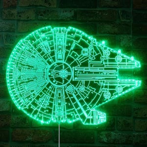 K&ouml;nnte beinhalten: Ein gr&uuml;nes Neon-Schild in Form des Raumschiffs Millennium Falcon. Das Schild leuchtet und ist an einer Backsteinmauer befestigt. Das Neon-Design zeigt die detaillierten Merkmale des Schiffs.
