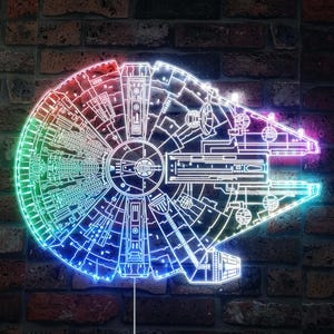 K&ouml;nnte beinhalten: Ein Neonschild, das das Raumschiff Millennium Falcon darstellt, mit detaillierten Umrissen und einem lebendigen, mehrfarbigen Leuchten. Das Schild ist an einer Backsteinmauer angebracht und zeigt ein Science-Fiction-Thema.