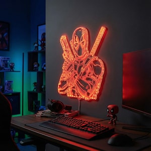 May include: A red illuminated Deadpool wall art piece. The artwork depicts Deadpool with two guns and two swords. The light is mounted on a dark gray wall. A computer, keyboard, and mouse are on a wooden desk below.