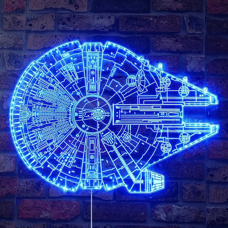 K&ouml;nnte beinhalten: Ein blaues Neonlicht in Form des Raumschiffs Millennium Falcon. Das Licht zeigt detaillierte Elemente des Schiffsdesigns und ist an einer Backsteinmauer befestigt. Das Licht leuchtet in einem leuchtenden Blau.