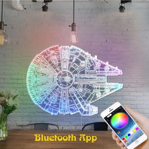 K&ouml;nnte beinhalten: Ein mehrfarbiges LED-Licht in Form des Raumschiffs Millennium Falcon. Das Licht ist an einer wei&szlig;en Backsteinmauer befestigt. Die Worte "Bluetooth App" werden in gelbem Text am unteren Rand des Bildes angezeigt. Ein Smartphone zeigt eine Farbrad-Oberfl&auml;che.