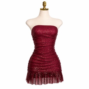 May include: A strapless, burgundy sequin dress displayed on a mannequin. The dress features a ruched bodice and a ruffled hem. The sequins catch the light, creating a shimmering effect. The dress is a mini-dress style.