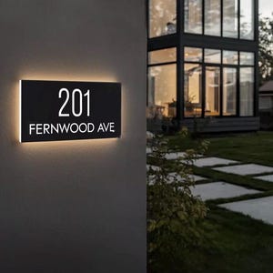以下が含まれることがあります： 黒い長方形の住所表示板。白い文字で「201」と「FERNWOOD AVE」と書かれています。温かく柔らかな光で照らされ、濃い灰色の壁に取り付けられています。