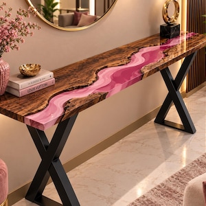 May include: A long console table featuring a natural wood slab top with a wavy, glossy pink resin inlay running through the center. The table is supported by two black metal X-shaped legs. Two books titled The Kinfolk and Louis Vuitton sit on the surface.