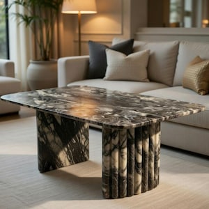 Op de afbeelding: Een rechthoekige salontafel met een marmeren blad met grijze en witte adering. De tafel heeft twee cilindrische poten met verticale groeven. De tafel staat in een woonkamer met een bank en een fauteuil.