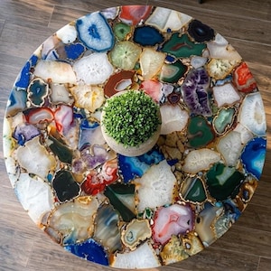 May include: A round coffee table with a mosaic top made of colorful agate slices. The agate pieces are in shades of blue, green, red, white, and purple, with gold-colored edges. A small potted plant sits on the table.