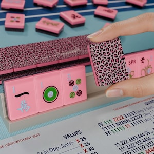 A hand is holding a pink pumkin with a leopard print pattern, with a pink and blue background featuring a game board and tiles.